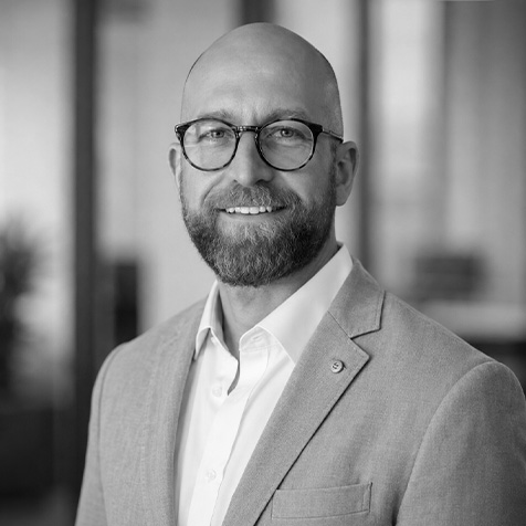 Black-and-white professional headshot of a person wearing round glasses, a light-coloured blazer and a white collared shirt. The person is facing the camera in an office setting, with a softly blurred background featuring glass panels and interior architectural elements. The portrait is cropped from the chest upwards and is evenly lit with a sharp focus on the subject.