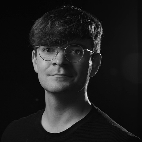Black-and-white studio portrait of a person wearing round glasses and a dark crew-neck top. The image is cropped from the shoulders upwards, with the subject facing the camera against a dark background. Dramatic side lighting creates strong contrast, highlighting facial features and hair while leaving parts of the background in shadow. The photograph has a professional, high-contrast style with sharp focus on the subject.