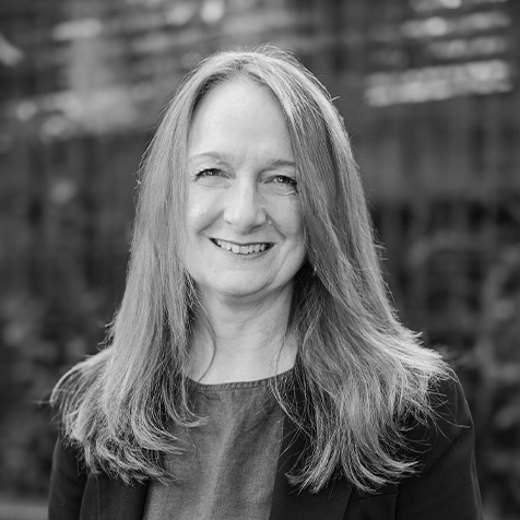 Black-and-white professional headshot of a person with long hair, wearing a dark blazer and top, standing outdoors against a softly blurred background.