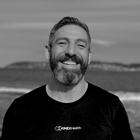 Black-and-white headshot of a person with short hair and a beard, standing outdoors in front of the sea, wearing a black KINEX Health T-shirt.